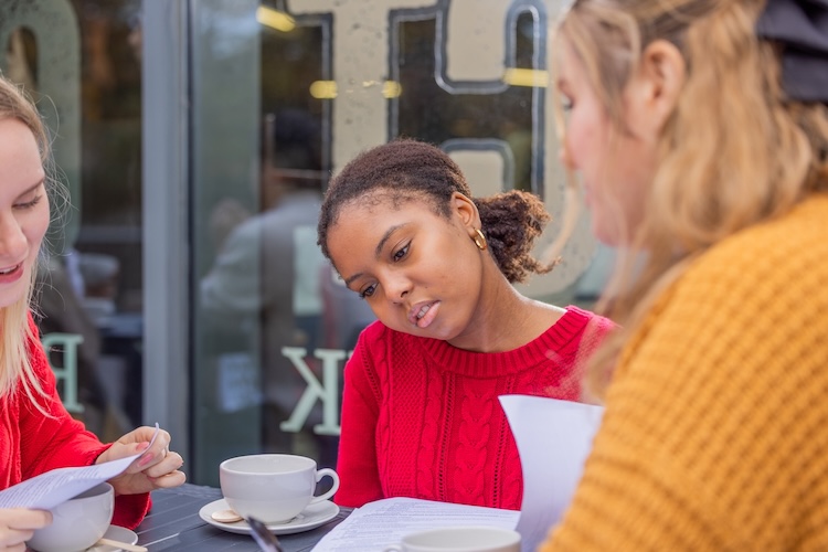 Three women sit at a café table, reading and discussing papers over cups of coffee.